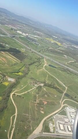 The video shows an aerial view of a green landscape with fields, roads, and some buildings under a clear blue sky. A highway is visible running through the landscape. The wing of a plane is partially visible at the bottom right corner. In the distance, mountains can be seen under the clear sky. The fields are varied, with some appearing green and others showing patches of yellow. There appear to be some residential buildings spread throughout. The overall impression is a view of a rural and suburban landscape from an airplane window.