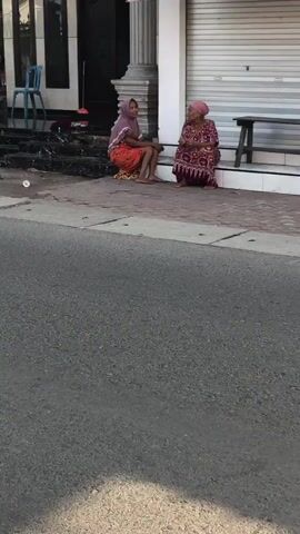 The video shows a street scene with several people and vehicles. In the foreground, a motorcycle is partially visible from behind, with its license plate in focus. The motorcycle is in motion, as indicated by the motion blur. Further in the background, a person is riding a red scooter, wearing a pink hijab and a mask. Another person, also wearing a pink hijab, is visible on the scooter behind the driver. The scooter appears to be moving on a paved road.

In the mid-ground, two women are sitting on steps outside a building. One woman, wearing a pink hijab and an orange and red patterned skirt, is sitting on the lower step. The other woman, wearing a patterned dress and a pink headscarf, is sitting on a higher step and appears to be engaged in conversation with the first woman. A blue plastic chair is placed near the steps.

The environment appears to be an urban or semi-urban area with a building that has pillars and closed metal shutters. The road is paved, and sunlight is casting shadows, suggesting it is daytime. The overall impression is that of a casual street scene in a community.
