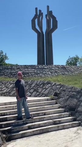 The video shows a tall concrete monument under a clear blue sky. In the foreground, a person with pink hair, wearing a dark t-shirt and light blue jeans, stands on a set of white stairs. To the right, another person in a beige shirt and grey shorts is seen taking a picture with their phone. The monument stands atop a hill covered with a stone wall and grass. A contrail from an airplane is visible in the sky.