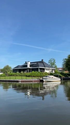 The video shows a house with a dark roof and light walls, situated on the edge of a body of water under a bright blue sky with scattered clouds.  A small boat is docked next to the house's property, reflected in the calm water. The lawn is green and well-maintained, with some trees and bushes surrounding the house. A flag is visible in the distance on the left side.