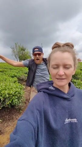 A woman in a blue hoodie with the word "Synchronicity" on it holds a camera and records herself and a man behind her. The woman is fair-skinned and has her blonde hair in a bun. She makes several faces for the camera while the man behind her is expressive and waves his arms. The man has a beard and is wearing sunglasses, a baseball cap, a gray t-shirt, and a black vest. They are in a field of green bushes, possibly tea plants, with a cloudy sky in the background. The camera is shaky.