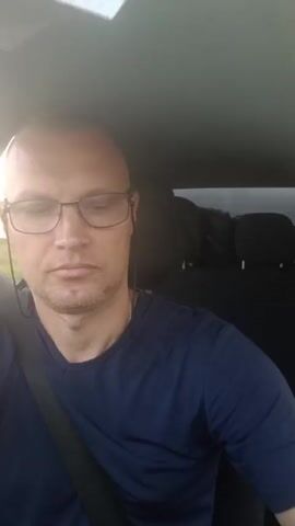 A man with glasses and a bald head is sitting in the driver's seat of a car, looking at the camera. He is wearing a dark blue t-shirt and a seatbelt. The sun is shining brightly through the window, creating a glare on the lens. The interior of the car is visible in the background, including headrests and what appears to be luggage. The man's expression is neutral.
