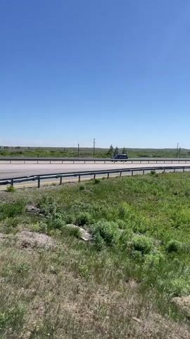 The video shows a highway on a clear, sunny day. The camera is positioned on a grassy embankment overlooking the road. A guardrail runs along the side of the highway. In the distance, the landscape is flat and grassy with power lines visible. A dark-colored car drives along the highway followed by a large semi-truck with "BTR" written on the side. More cars are seen further down the road. A weathered wooden fence stretches along the grassy area.
