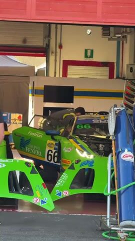 A race car pit crew works on a green and yellow race car with the number 66 visible. The car's hood is open, and one mechanic is leaning into the engine bay. A man in a black t-shirt and jeans stands observing the work. The scene takes place inside a garage with red garage doors and yellow and blue striped walls. Another car, also green and yellow, is visible further back in the garage, and there are various pieces of equipment around.
