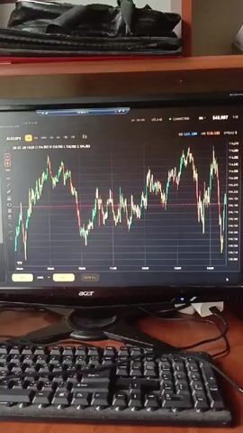 The video shows a close-up shot of a computer monitor displaying a financial trading chart. The chart shows price movements of a currency pair, likely AUD/JPY, with candlesticks indicating price fluctuations over time. The screen also shows various trading indicators and data, such as current price, spread, and connection status. A black computer keyboard is visible in the foreground, positioned in front of the monitor. The background is slightly blurred, but appears to be a room with a wooden shelf or desk and some dark fabric objects. The lighting suggests an indoor environment. The video is very short, showing only a few seconds of the trading chart.