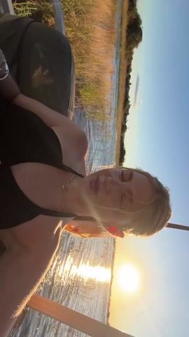 A woman with blonde hair smiles while riding on a small boat, the sun low in the sky behind her. The woman is wearing a black top and a silver necklace. She has a scarf tied in her hair. She appears relaxed, looking around at the scenery as the boat moves through the water. Tall grass grows along the shoreline. The sky is clear and blue.