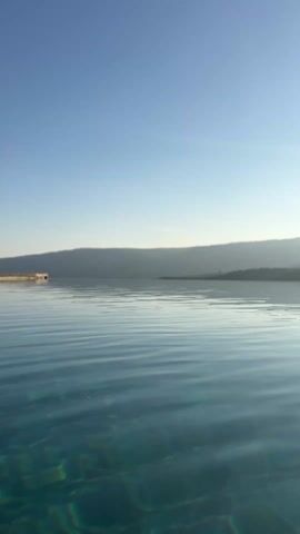 The video shows a wide, tranquil body of water under a clear, pale blue sky. In the distance, a blurry, low-lying landmass stretches across the horizon. In the foreground, the water exhibits subtle ripples, creating a pattern of light and shadow. The water's surface is clear enough to reveal a textured bottom underneath. On the left side of the frame, a small, concrete structure or platform is visible near the water's edge.