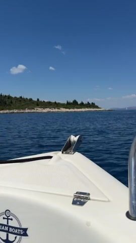 The video shows the bow of a white boat moving through blue water under a clear blue sky with scattered clouds. The boat has a metal anchor roller and a hinge visible on its deck. In the background, a green, tree-covered island or coastline is visible on the horizon. The logo of "DALMATIAN BOATS" with an anchor is visible on the side of the boat. The camera is positioned looking forward from the boat.