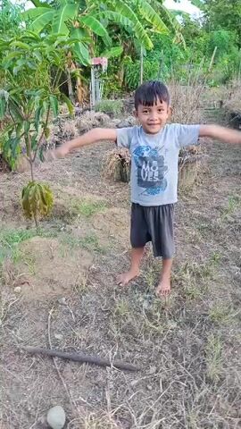 A young boy stands in a dirt yard with dry grass and some plants. He is wearing a gray t-shirt with a graphic on it and dark shorts. His arms are extended out to his sides and his hands are pointed downwards. He is looking at the camera and smiling. In the background, there are trees and some structures.