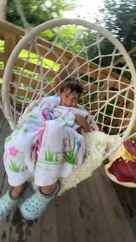A young child is sitting in a hanging chair, wrapped in a white towel with a floral pattern. The child has dark, wet hair and is looking down. The hanging chair is made of rope and is suspended from a wooden structure. The background shows trees and greenery. The child appears to be moving or being moved, as the camera is slightly shaky. The child is smiling and looking at the camera at the beginning of the clip.