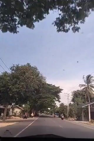 The video shows a view from the inside of a car driving on a road. The sky is blue with a few scattered clouds. The road is asphalt and has painted white lines on the sides. Large, green trees line the sides of the road, creating a canopy overhead in some sections. Several power lines are visible running alongside the road. There are some buildings on the sides of the road, and a couple of people walking along the shoulder. A few motorbikes are also seen driving in the same direction as the car. The general environment suggests a rural or suburban area.