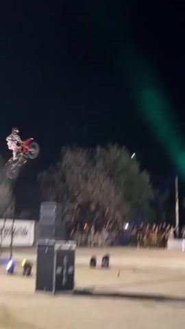 A motocross rider in a red and white suit performs a trick mid-air against a dark night sky. A beam of light, possibly from a spotlight, illuminates the rider and extends downwards, creating a smoky trail. Below, a crowd of blurred figures watches from behind barriers, with trees and buildings visible in the background. In a subsequent shot, the rider is seen at the peak of their jump, silhouetted against the dark sky. The video then cuts to a ramp structure, followed by shots of the ramp from a different angle, with a crowd gathered and lights illuminating the area. Finally, a man in a dark t-shirt and shorts is shown speaking into a microphone, with a ramp and a crowd in the background, suggesting he is an announcer or presenter.