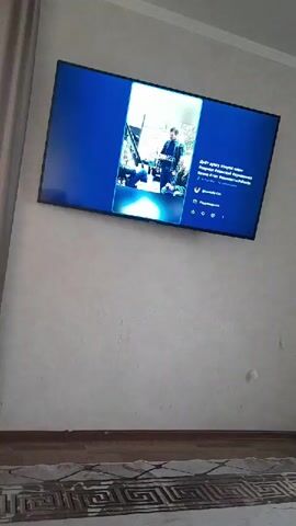 A wall-mounted television displays a social media post featuring two individuals. The environment includes a wall with a curtain to the left and a patterned rug on the floor beneath the television. The social media post on the screen has a predominantly blue interface.