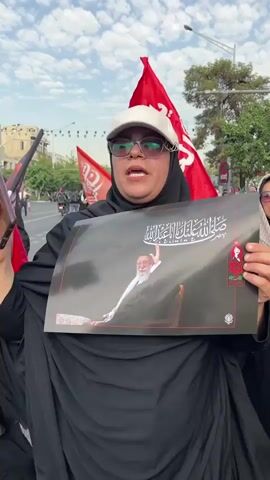 A medium shot captures a woman in the foreground, wearing a black hijab and abaya, with a white baseball cap and sunglasses. She is holding up a large poster with an image of an elderly man with a white beard and a black turban, seated and gesturing with his right hand. Arabic script is visible on the poster. She is also holding a mobile phone in her left hand. Behind her, several red flags with Arabic script are displayed, along with more individuals in the background, some of whom are also holding posters and flags. The scene appears to be outdoors, with trees and a street visible. The overall atmosphere suggests a rally or protest.