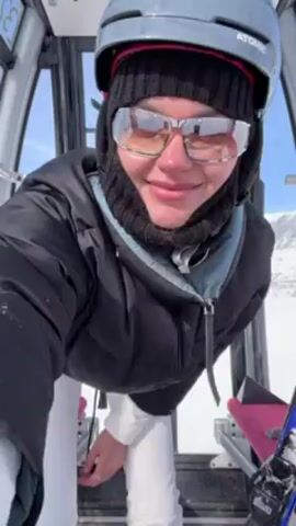 Inside a ski lift, a person wearing a helmet, goggles, a black balaclava, a black puffer jacket, and white ski pants is dancing. The person is facing the camera initially, and then turns around to face the window of the ski lift, where a snowy mountain landscape is visible. The ski lift has red seats and a grey frame.