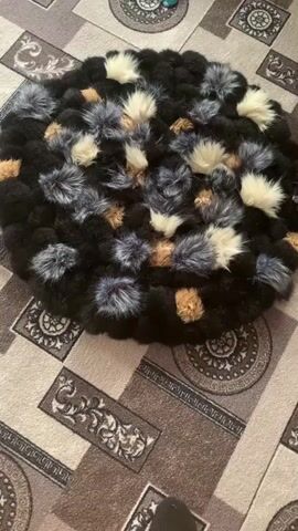 A black, circular rug with patches of white, brown, and grey faux fur is displayed on a tan carpet with a geometric pattern. The rug is covered in a dense arrangement of black fluffy material, interspersed with smaller, pom-pom-like tufts of faux fur in white, tan, and grey. The camera is positioned above and to the side of the rug, slowly rotating around it, showcasing the texture and color variations of the rug.