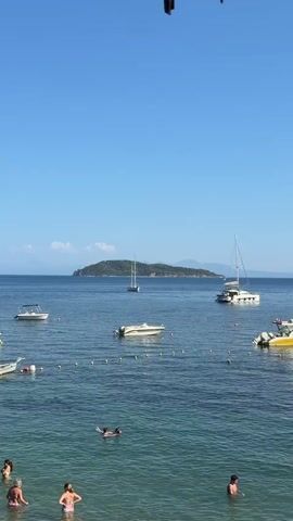 The media file depicts a serene scene at a beach in Skiathos, Greece. Several boats are anchored in the clear blue water, including sailboats, a catamaran, and smaller motorboats. Some people are swimming and wading in the shallow water, while others are enjoying the sun and sea from the boats. The sky is a vibrant, cloudless blue, suggesting a warm, sunny day. In the distance, a lush green island with rolling hills stands against the horizon, adding to the picturesque setting. The water is calm and inviting, with gentle ripples on its surface. The overall mood is one of relaxation and enjoyment, indicative of a perfect summer day at a Greek island resort. There is no visible text in the media file.