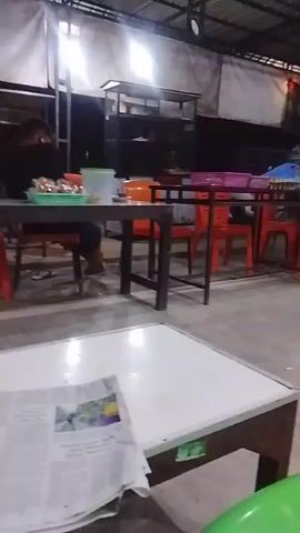 The media file depicts an indoor or semi-outdoor seating area, likely a restaurant or food stall, at night. The scene is dimly lit, with artificial lights casting a warm glow on the tables and chairs. Several tables are visible, some with orange chairs. In the background, a screen, possibly a television, is displaying content, and there are shelves stocked with various items.

There are at least two individuals present. One person, wearing a light blue shirt, is seated at a table, seemingly engaged in eating or conversing. Another person is partially visible on the right side of the frame, also seated at a table. The overall atmosphere appears relaxed, suggesting a casual dining experience. The floor is concrete, and the ceiling is covered with a corrugated metal roof supported by metal beams, common in many Indonesian food establishments. A newspaper is visible on a white-topped table in the foreground. The location is identified as Dayahlhoksukon Barat, Indonesia.