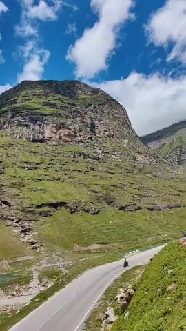 The video pans across a mountainous landscape. A paved road winds through the valley, with a lone motorcyclist visible on the road. Green slopes covered in vegetation rise steeply on either side of the valley, with rocky outcrops and cliffs visible. A few small structures and patches of farmland are present in the valley floor. The sky above is blue with scattered white clouds.