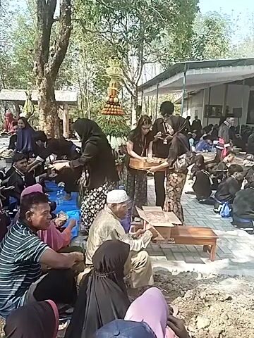 The video shows an outdoor gathering, possibly a celebration or ritual, in a rural or semi-rural setting. Several people are seated on the ground, some on blue tarps, in what appears to be a communal meal or distribution of food. In the center, a tiered display of fruits and vegetables is prominently featured, resembling a decorative centerpiece or an offering. A group of individuals, some wearing traditional clothing, are interacting around this display, with some appearing to be serving or distributing items from it. There are more people seated in the background, and a building or structure is visible behind them, suggesting an organized event. The overall atmosphere seems to be one of community participation and shared activity.