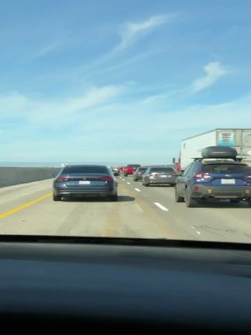 A blue sedan drives on a highway in moderate traffic. The sky is blue with wispy clouds. The car is in the right lane, with several other cars and a semi-truck in front of it. The license plate of the blue sedan reads "9MTULL6". The camera appears to be mounted inside the car, looking out the windshield.