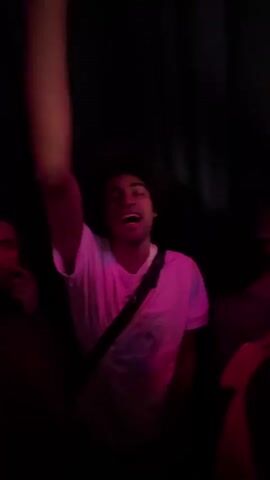 The video shows a group of people in a dark environment, possibly a club or concert, illuminated by red and pink lighting. Initially, a man with dark hair and a white shirt is visible with his arm raised. The camera then focuses on a young woman with blonde hair, who is shouting or singing along with the music, displaying visible excitement. The scene is energetic and dynamic, with people moving and reacting to the music.
