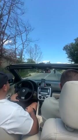 Two men are in a black convertible Volkswagen Eos, driving down a paved road on a sunny day. The driver, wearing a black baseball cap and a white t-shirt, is holding the steering wheel. The passenger, in the right seat, is wearing a light-colored t-shirt. The top is down, and the sky is blue with scattered clouds. The road is flanked by trees and greenery. There is a motorcycle visible in front of the car, and another car in the distance. The car's interior is visible, including the dashboard and center console with a smartphone mounted on a holder. The video is shot from the perspective of someone in the back seat, looking forward.
