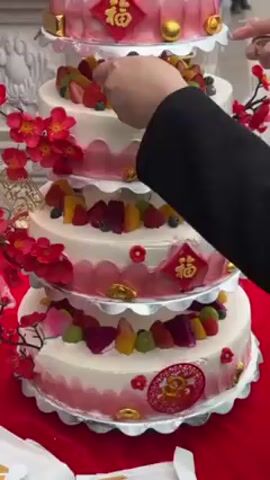 A person is carefully arranging the tiers of a five-layer cake outside in front of a building with columns. The cake is decorated with white and pink icing, red and gold Chinese characters, various fruits, gold spheres, and sprigs of red flowers. The cake tiers are separated by silver platforms.  The person, wearing a black jacket, adjusts the position of the tiers, adding more support.  Other people are visible in the background, moving about near the building. A red cloth drapes across the surface the cake is being assembled on.