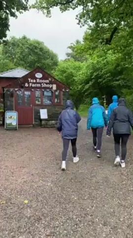 Here is a detailed description of the video:

**Scene:** The video shows a group of people walking towards a Tea Room & Farm Shop on a rainy day.

**Environment:** The setting is a lush, green area with many trees. It appears to be a park or nature area. The ground is gravelly. It's raining, as indicated by the wet ground and the people's attire.

**People:** There are at least four people visible in the video. They are wearing rain jackets and leggings/trousers, suggesting they are dressed for wet weather. They are walking together towards the Tea Room & Farm Shop.

**Objects:** 
*   **Tea Room & Farm Shop:** A small building with a red door and a grey stone base. There are signs and posters outside.
*   **Signs:** There are signs posted near the building.
*   **Picnic Table:** There is a picnic table in front of the building.
*   **Wooden Fence:** A wooden fence borders a part of the area, suggesting a boundary or edge to the park.
*  **Signage:** There is a small sign with a picture in front of the tea room.

**Actions:** The people are walking towards the Tea Room & Farm Shop. The camera pans and zooms slightly to capture different parts of the scene.

**Interactions:** The group of people are walking together, possibly intending to visit the tea room.