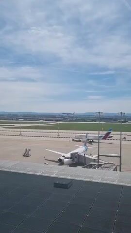 The video shows an airport tarmac on a sunny day. In the foreground, a white airplane with blue trim is parked, connected to a jet bridge. A set of stairs is also positioned nearby. In the middle distance, another white plane is visible near what appears to be ground crew. Further away, a blue KLM airplane is landing on a runway, with a city and hills in the background. The camera pans slightly, and airport vehicles are visible.