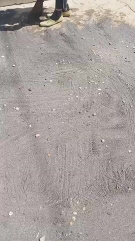 Here's a description of the video:

The video shows someone sweeping a section of paved ground that is covered in fine gray gravel or sand. There are small rocks and debris mixed in with the sand. The person uses a straw broom, and the sweeping motion creates swirling patterns in the material. The edge of the pavement is visible, contrasting slightly in color. At one point, the shadow of the person holding the camera falls on the ground.  A drain cover is located near one edge of the paved area.