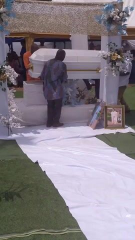 The video shows three men carrying a white casket across a green, astroturf covered outdoor space, likely during a funeral. The casket is trimmed with gold accents. A white runner lies on the astroturf, and framed photographs are propped against a white structure decorated with white and blue flowers. White tents cover the scene, and other people can be seen in the background.