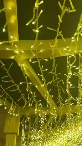 The video shows a structure decorated with strings of small, warm-white LED lights hanging from a wooden frame. The lights create a canopy effect, densely packed and glowing brightly. The structure has support columns, and the lighting extends downwards, creating a tunnel-like effect. The environment appears to be outdoors at night, with the darkness surrounding the illuminated structure. A voice of a child can be heard.