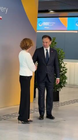 The video shows two individuals, a woman and a man, shaking hands. The woman is on the left, wearing a white textured blazer over a white top and black wide-leg pants. She has curly, light brown hair and is wearing glasses. The man is on the right, dressed in a dark navy suit, a light blue tie, and a white shirt. He has short brown hair and is also wearing glasses. They appear to be in a professional setting, with a backdrop featuring a large banner.

The banner displays the text "Ukraine Recovery Conference" in English, with the dates "25-26 June 2023" and the location "Gdańsk, Poland" below it. The logo for the conference, featuring a circular orange and blue design with abstract shapes, is also visible. To the right of the banner, there is a large potted green plant.

The lighting suggests it is daytime, possibly indoors in a well-lit conference or event space. The interaction appears formal and polite, with both individuals smiling. The overall scene is one of a professional meeting or exchange at an international conference focused on the recovery of Ukraine.