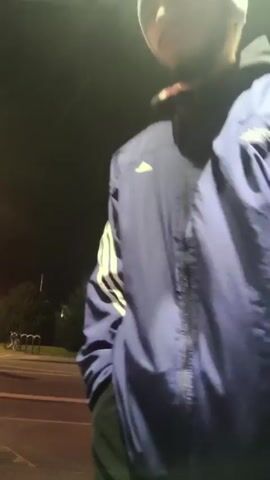 A man wearing a blue tracksuit and a grey beanie is seen on a street at night. He gestures towards the camera, then throws the object being held. The camera is then pointed towards a building across the street with a lit-up sign. The surrounding environment includes trees, a streetlight, and a bicycle rack.
