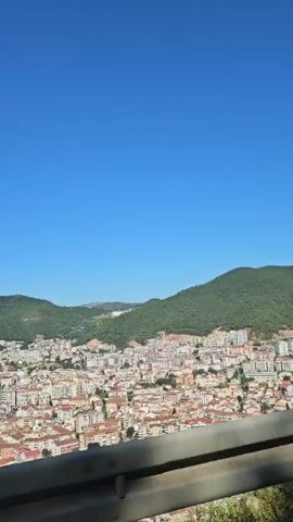 A long, vertical shot begins with a view of a coastal city nestled between green, tree-covered hills and a bright blue, clear sky. A white barrier runs horizontally across the bottom of the frame, obscuring the foreground. As the camera moves, the city expands into view, revealing numerous buildings with terracotta-colored roofs and white walls, densely packed together. A harbor filled with boats is visible on the left side, and the turquoise water of the sea stretches to the horizon. The camera continues to pan right, showcasing more of the urban sprawl and the curving coastline. Sunlight illuminates the scene, creating a bright and airy atmosphere. The video ends with a view of the city and the sea under a vast, cloudless blue sky.