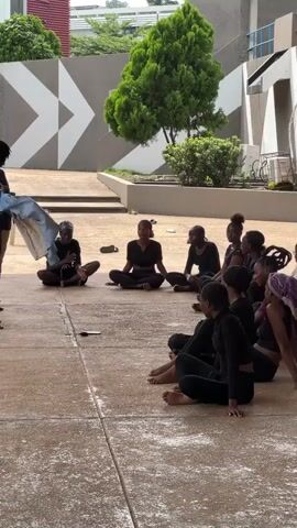 In an outdoor courtyard, a group of young Black women, dressed in black athletic wear and some with their hair tied back, are seated in a circle on the ground. One woman stands in the center of the circle, holding a blue garment, and appears to be demonstrating something to the seated group. Another woman, also dressed in black, stands to the side of the central demonstrator. The ground is paved with concrete tiles. In the background, a building with a gray facade features a large geometric pattern in white. Lush green trees are visible against the building and in planters. The time is daytime.