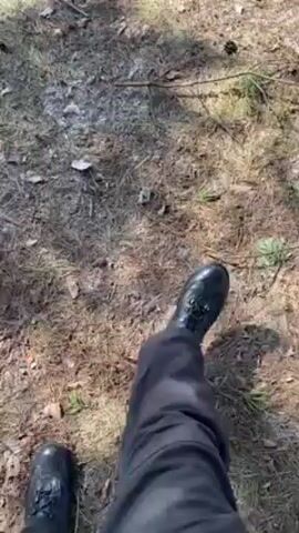 The video begins with a low-angle shot of the ground covered in pine needles and sparse grass. A person in black boots and pants walks across the surface. As the camera moves, it reveals a view of a snowy landscape framed by bare trees. There is a partially frozen lake or pond visible in the background. The sky is pale blue.