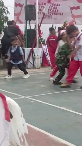 A group of children are participating in an outdoor event on a court marked with white lines. One child in the foreground, wearing a light blue shirt and dark pants, moves towards the right of the frame, looking back. Another child, dressed in a green camouflage military uniform, is captured in motion, appearing to be mid-stride or jump. Further back, several other children are visible, some in red outfits. Adults are also present, with one in a red shirt standing near a speaker on a raised platform. A banner with red and white patterns, and text that appears to be in Indonesian, is displayed prominently in the background, suggesting a national celebration. The ground is a reddish-brown asphalt with green sections. The overall atmosphere seems festive.