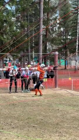 In an eye-level, outdoor shot, four people participate in an obstacle course involving ropes.  

The individuals, dressed in climbing gear including harnesses and helmets, appear to be a team. One person wears orange clothing, another white with a blue beanie, another white with a yellow helmet, and the last in white with a white helmet. They stand near a tall, cylindrical, striped pole with multiple ropes strung from it to the surrounding trees. They are holding ropes and adjusting their gear.  

Behind the participants is a red mesh barrier, and behind the barrier is a group of spectators in casual clothing. Tall pine trees surround the area. The ground is a mix of dry grass and patches of dirt.  

The video appears to capture a moment of preparation or instruction before attempting the rope obstacle.