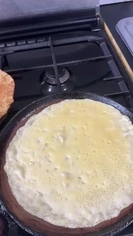 The video shows a close-up shot of crepes being cooked on a stovetop. A black cast-iron pan is placed on a black gas stove. A pale yellow liquid, likely crepe batter, is being poured into the pan. As the batter cooks, it forms small bubbles on the surface, indicating it is setting. A spatula is used to lift one edge of the cooked crepe, revealing a golden-brown underside. Another crepe, already cooked and golden-brown, is visible to the left of the pan. The video captures the process of making crepes, from pouring the batter to flipping the cooked crepes.