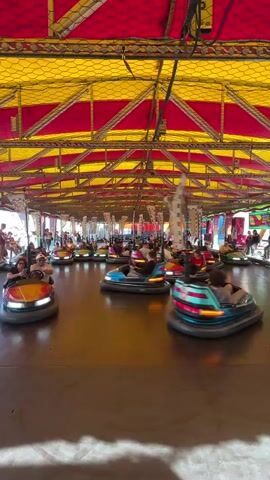 The video captures an energetic scene inside a bumper car arena. Numerous individuals, including adults and children, are seen actively driving and bumping colorful bumper cars (red, blue, yellow, and orange) across a smooth, dark floor. The arena is covered by a distinctive red and yellow striped canopy or tent, supported by a visible metal framework and netting that partially encloses the space. Overhead, poles extend from the ceiling to connect with the individual bumper cars, providing power.

The atmosphere appears lively and fun, typical of an amusement park or fairground setting. Some cars are in motion, colliding with others, while a few are temporarily stationary. Bright light suggests it is daytime. No specific text or signs are clearly visible within the frame. The overall impression is one of recreational activity and enjoyment.