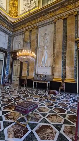 The video pans across a grand, opulent room with a high ceiling. The walls are adorned with intricate bas-relief sculptures, featuring classical figures and architectural elements, framed by gilded columns. A large, ornate crystal chandelier hangs from the center of the ceiling, casting a warm glow. The floor is a striking mosaic of geometric patterns in contrasting colors. In the foreground, several small, upholstered stools with ornate golden legs are placed around the room. In the middle ground, a long, narrow table with decorative accents stands against the wall, topped with a vase and candelabra. The overall impression is one of historical grandeur and luxury. No people are clearly visible in the frame.