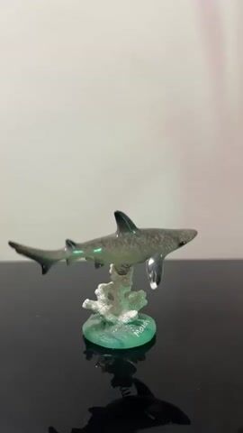 This media file features a close-up shot of a small, decorative shark figurine. The figurine is made of glass and is depicted swimming above a piece of white coral, which is attached to a translucent green base. The shark is grey with darker stripes and has a shiny finish, reflecting the light. The base of the figurine has the word "Malé" etched onto it in a light green script. The figurine is placed on a dark, reflective surface, possibly a table, which shows a distorted reflection of the shark. The background is a plain, light-colored wall, slightly out of focus. The lighting appears to be artificial, casting a subtle glow on the figurine. There is no discernible activity or event taking place, as it is a still object. The setting appears to be indoors, likely a display or shelf. The text "Malé" suggests a possible connection to Malé, the capital city of the Maldives, hinting at a potential origin or theme for the souvenir.