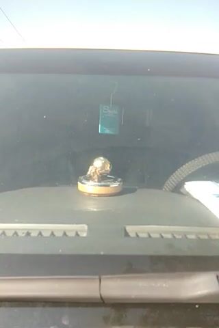The video shows an interior view through what appears to be a vehicle's windshield. Visible through the windshield is a light bulb, illuminated, mounted on a circular base. To the left of the bulb, a small rectangular object, possibly an air freshener, is hanging from the windshield with a string. The view is slightly obscured by the glass, and the environment outside is not clearly visible due to the bright light. The camera is static, and the view is angled slightly downward.