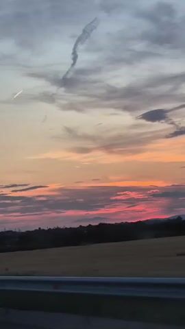 This media file captures a picturesque sunset over a rural landscape in Yana, Bulgaria. The sky is a canvas of vibrant colors, transitioning from a soft gray at the top to warm hues of orange, pink, and red near the horizon, indicative of a late evening. Scattered clouds create texture and depth, with a distinct contrail forming a wispy line across the upper portion of the sky.

The foreground features a dark silhouette of trees and foliage against the brightly lit sky, suggesting a transition from day to night. Below the treeline, a field appears to be harvested or in a state of late growth, its golden-brown color hinting at a dry season. A metallic barrier, possibly part of a road or viewing platform, runs across the bottom of the frame, framing the scene.

The overall mood conveyed is one of tranquility and natural beauty. The absence of people or specific objects of human activity, aside from the barrier, emphasizes the serene, natural setting. The time of day is clearly sunset, with the vivid colors and fading light providing a peaceful ambiance. No visible text is present in the media file.