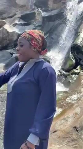 A woman wearing a navy blue long-sleeved shirt and a patterned headscarf stands near a waterfall. The waterfall cascades over gray rocks in the background. The woman adjusts her headscarf and looks directly at the camera.