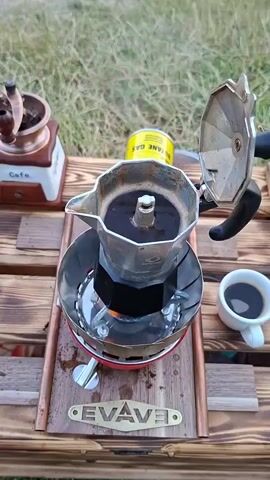 A close-up overhead shot shows a stovetop coffee maker with espresso brewing. The coffee maker sits on a portable stove with flames visible beneath it. A small white cup of coffee is to the right of the coffee maker, and a hand-crank coffee grinder is in the upper left corner. A can of butane gas is also visible in the background, along with a wooden tray. The scene appears to be outdoors, with grass in the background.