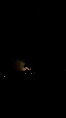 The video displays a dark, nighttime scene. The sky is entirely black, with no visible stars or moon. In the center-midground, a distinct plume of light brown or orange-tinged smoke is visible, illuminated from below by an unseen light source, suggesting a fire or burning activity. Below this smoke, scattered across the lower portion of the frame, are several small, out-of-focus, blue-white lights. These distant lights likely belong to buildings, streetlights, or other artificial sources in what appears to be a settlement or area with some infrastructure. There are no people visible in the shot, nor are there any clearly identifiable structures or objects beyond the smoke and lights. The overall atmosphere is dark, with the illuminated smoke providing the main focal point against the deep black. The audio includes faint human voices, suggesting people are present nearby, but not in the frame. The scene is set outdoors at night, with contextual information placing the location in Igbara, Nigeria.
