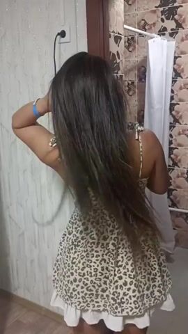 The media file shows a woman with long, dark hair from behind. She is wearing a short, sleeveless dress with a leopard print pattern and a white ruffled hem. The woman is in a room that appears to be a bathroom, indicated by the presence of a shower curtain and tiled walls. She is holding a black hairdryer in her right hand and is using it to dry her hair. Her left arm is raised, and she is using her hand to style her hair as she dries it. The lighting suggests it is daytime. There are no visible signs or text in the media file. The location context provided is Шұбартүбек, Kazakhstan.